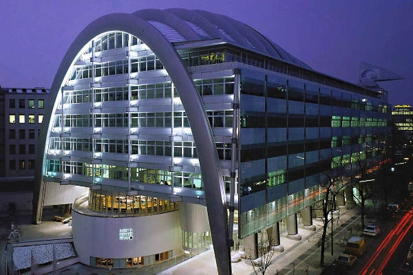Modern office building with striking arch-shaped architecture at dusk, large glass façade with illuminated interiors, urban street with traffic trails in the foreground. Ludwig Erhard Haus, communication center of the Chamber of Industry and Commerce in Berlin.