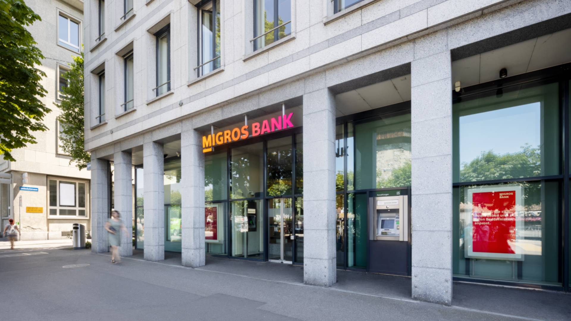 Aussenansicht einer Migros Bank Filiale mit moderner Steinfassade, grossen Glasfronten und leuchtendem Logo; Passanten vor dem Eingang und Bancomat sichtbar, Symbol für Banking, Customer Experience und Customer Relationship Management mit der BSI Customer Suite
