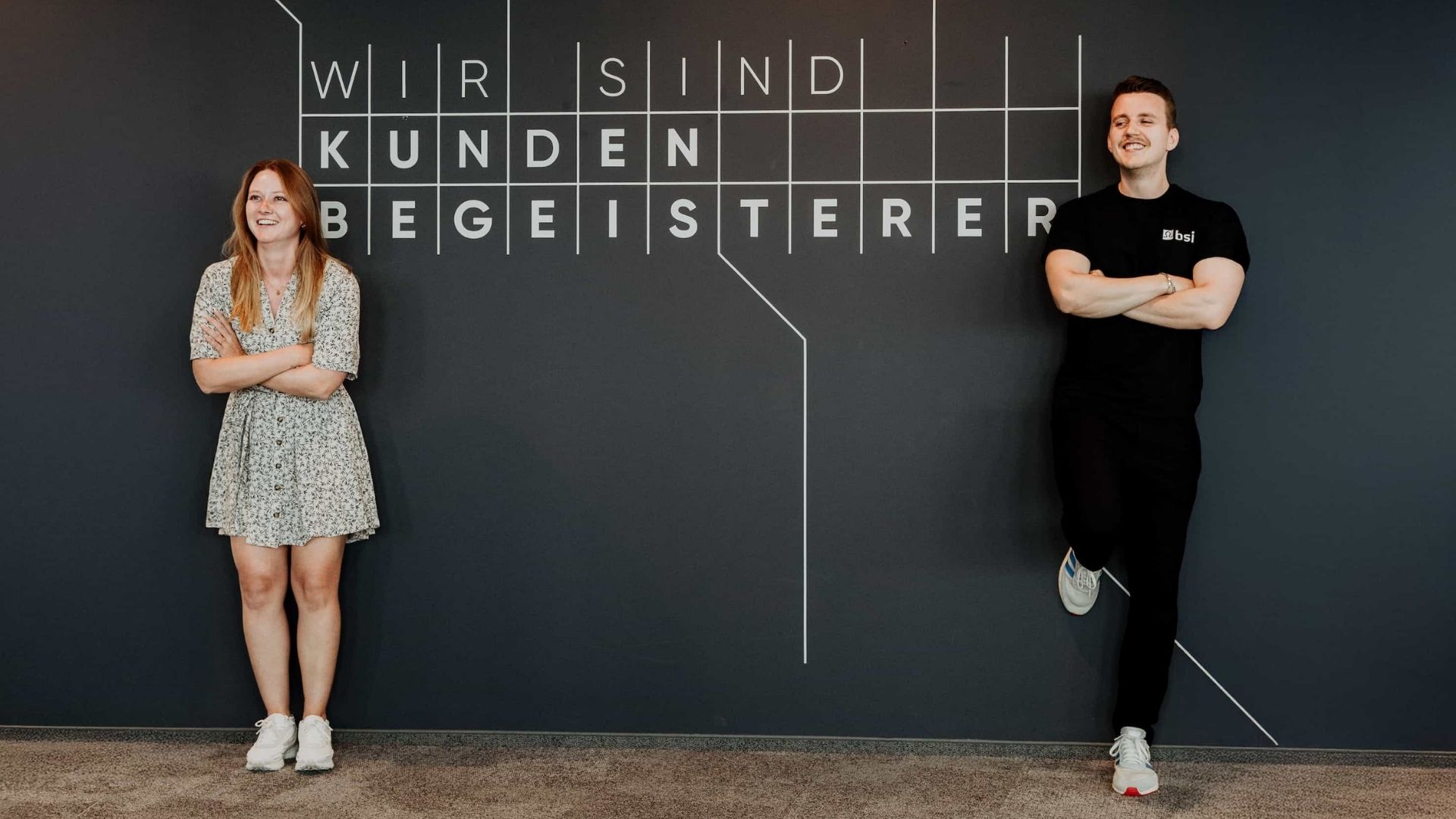 Zwei Mitarbeitende stehen lächelnd vor einer dunkelgrauen Wand mit der Aufschrift «Wir sind Kundenbegeisterer». Links steht eine Frau in einem hellen geblümten Kleid, rechts ein Mann in schwarzer Kleidung mit verschränkten Armen; modernes Büroambiente und freundliche Teamatmosphäre.