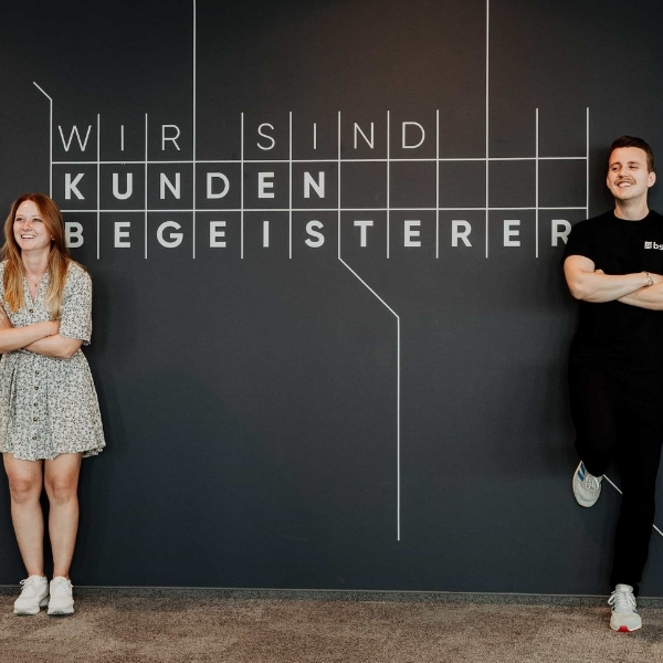 Zwei Mitarbeitende stehen lächelnd vor einer dunkelgrauen Wand mit der Aufschrift «Wir sind Kundenbegeisterer». Links steht eine Frau in einem hellen geblümten Kleid, rechts ein Mann in schwarzer Kleidung mit verschränkten Armen; modernes Büroambiente und freundliche Teamatmosphäre.