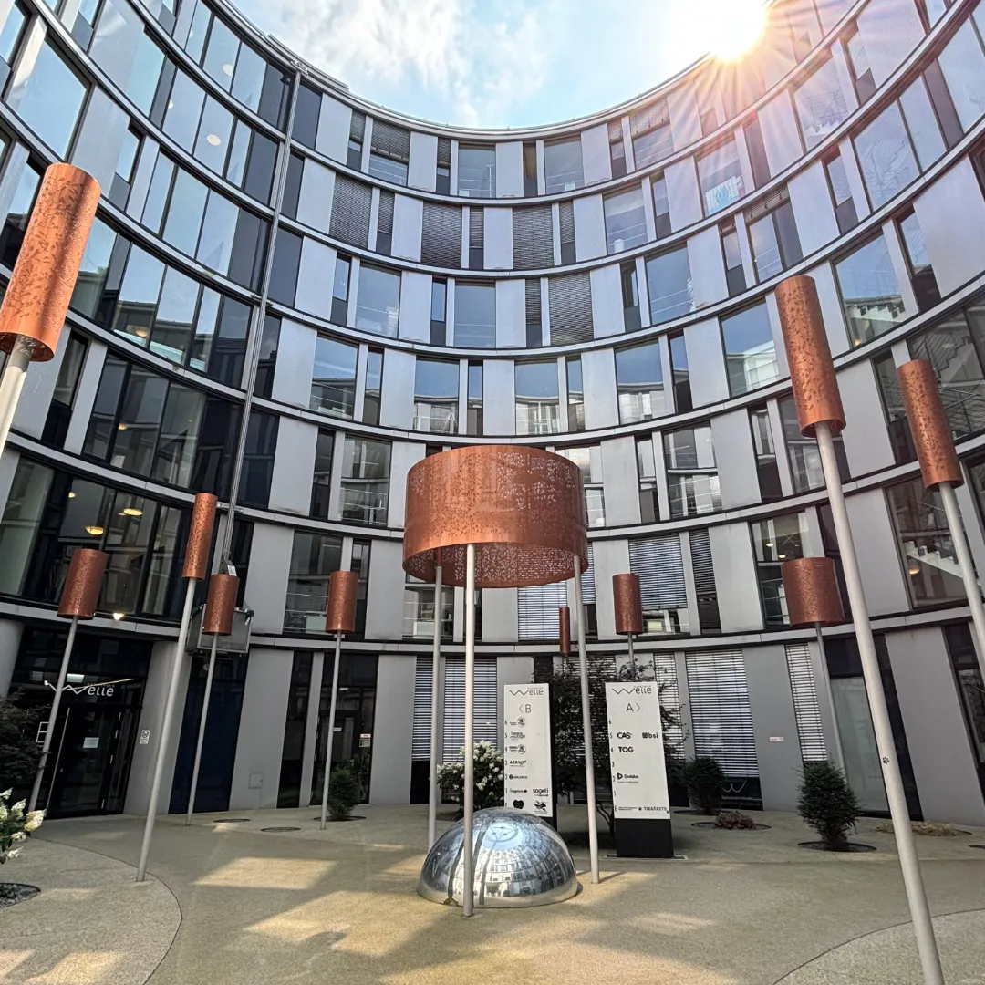 Sonnendurchfluteter Innenhof am BSI Standort Hamburg mit moderner, geschwungener Glas- und Metallfassade. Rostfarbene, zylindrische Leuchten stehen rund um eine spiegelnde Halbkugel; auf der Gebäudestele ist das BSI-Logo zu sehen.