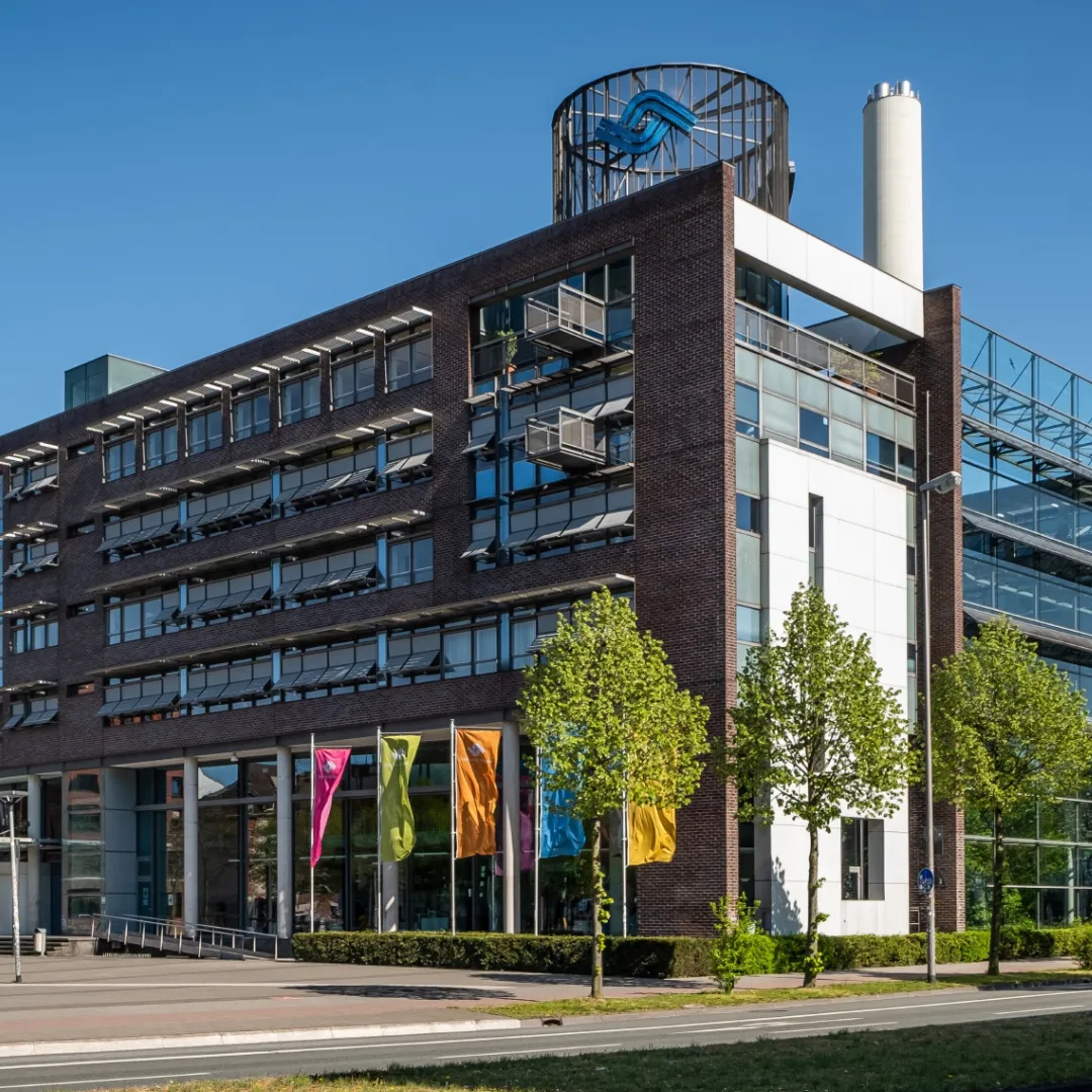 Gebäude am Hafenplatz in Münster, Sitz der Stadtwerke Münster. Das moderne Backsteingebäude mit grossen Fensterfronten, farbigen Fahnen und dem Stadtwerke-Logo auf dem Dach steht an einer begrünten Strasse. Stadtwerke Münster ist Kunde von BSI Software.