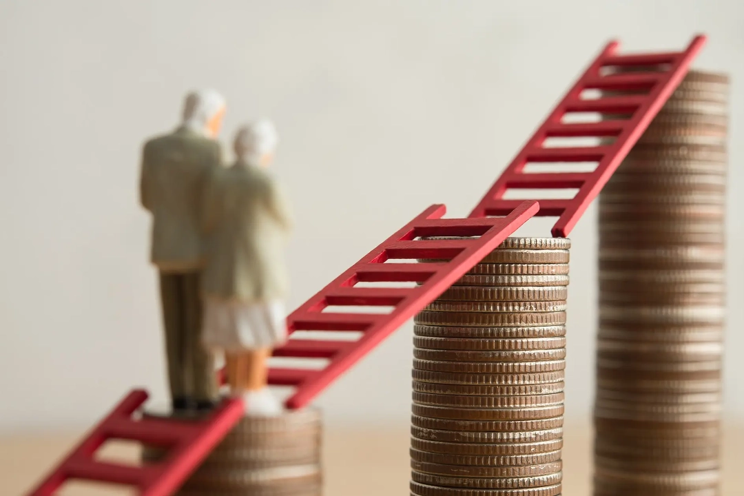 Zwei Miniaturfiguren eines älteren Paares stehen auf einem Münzstapel. Rote Leitern verbinden mehrere unterschiedlich hohe Münzstapel und symbolisieren den langfristigen Vermögensaufbau für die Altersvorsorge. Das Titelbild illustriert ein Interview mit BSI Software Vorstandsmitglied Burkhard Oppenberg über die Chancen des Altersvorsorgedepots für Versicherer.