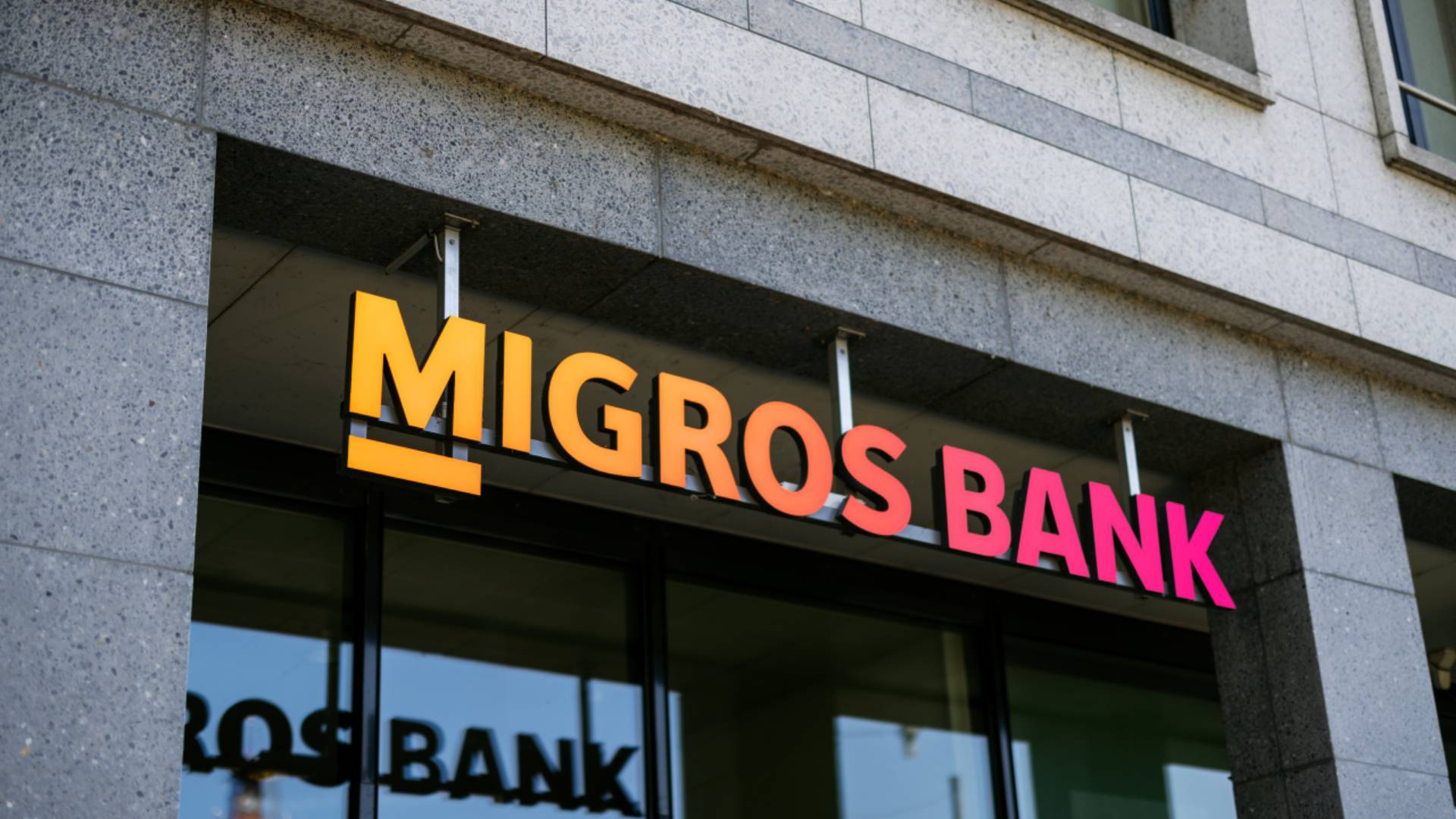 Aussenansicht einer Migros Bank Filiale mit leuchtendem Logo an moderner Gebäudefassade; Symbol für Banking, Customer Experience und Customer Relationship Management mit der BSI Customer Suite