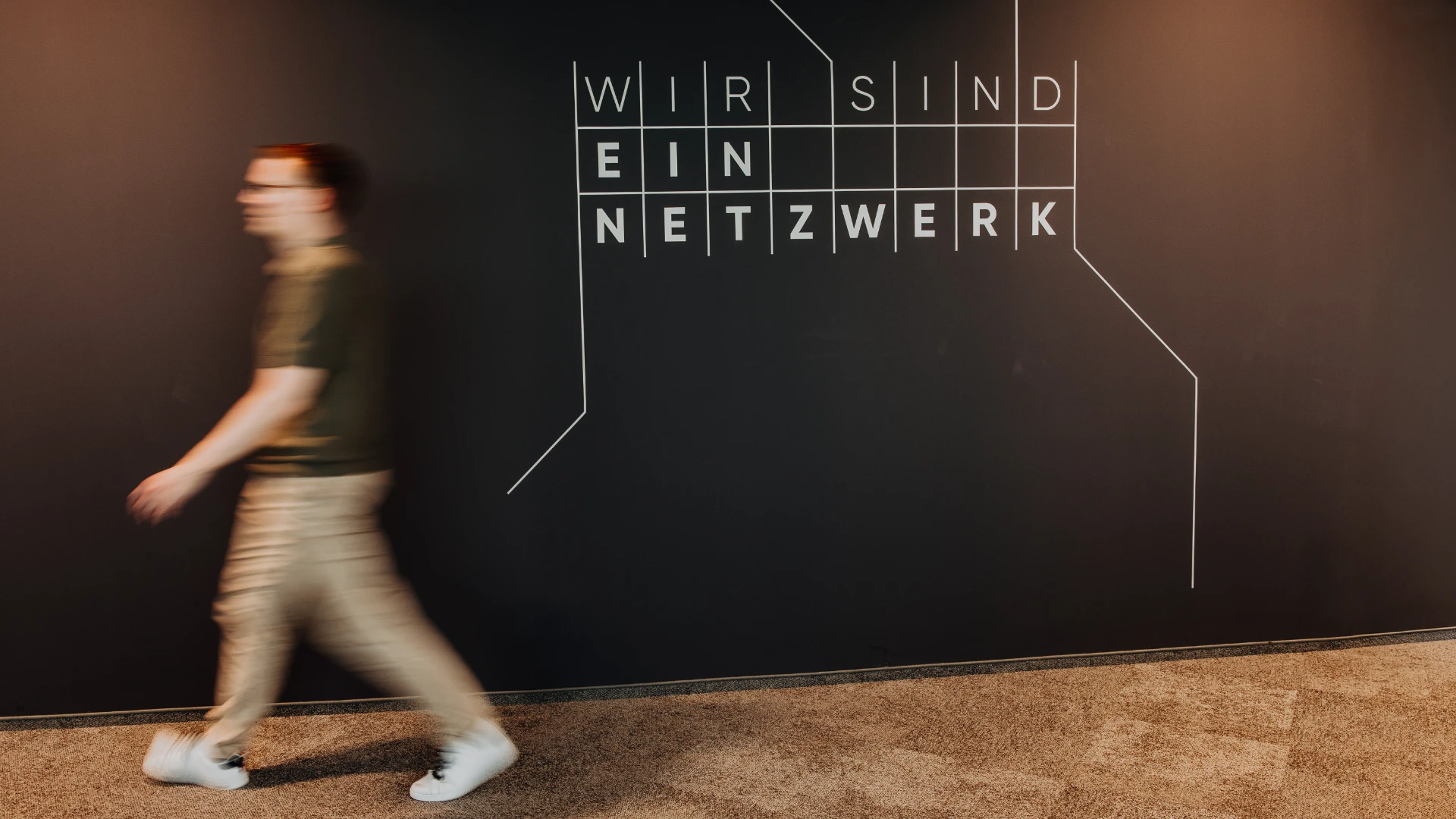 Mitarbeitender geht an einer Wand mit der Aufschrift «Wir sind ein Netzwerk» vorbei – modernes Office-Design und gelebte Vernetzung spiegeln die offene Unternehmenskultur und attraktive Arbeitsumgebung bei BSI Software wider.