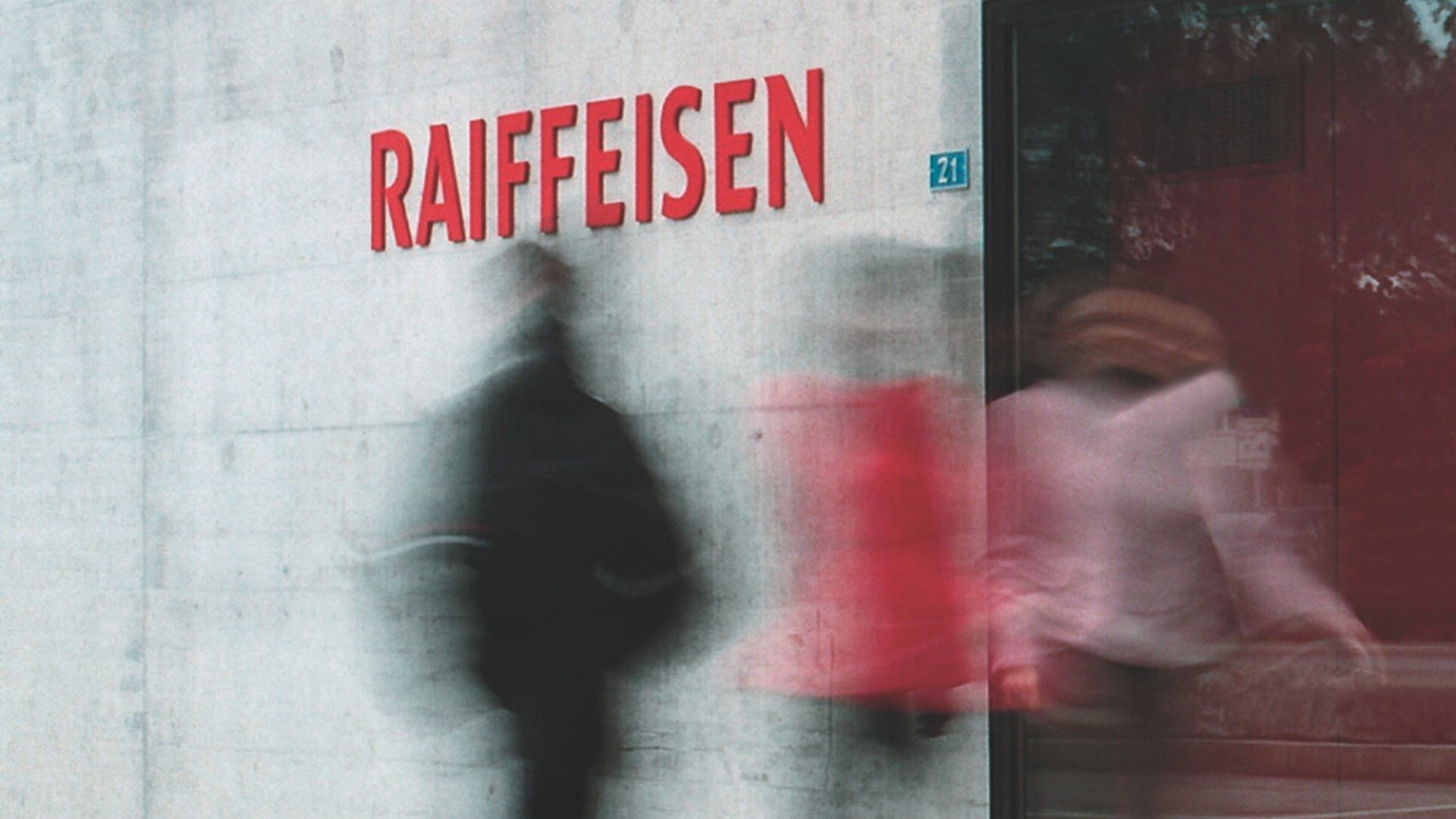 Motion blur of two people walking past the facade of a Raiffeisen bank branch with the red “RAIFFEISEN” sign.