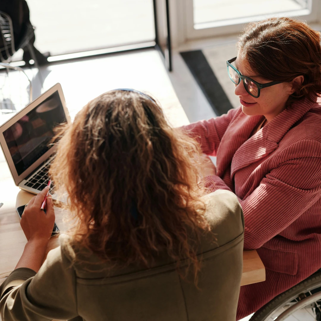 Zwei Frauen in einer persönlichen Beratungssituation im Versicherungskontext. Eine Versicherungsberaterin mit Brille und rosa Pullover bespricht mit einer Kundin am Laptop mögliche Versicherungslösungen. Die freundliche Gesprächsatmosphäre unterstreicht die Bedeutung individueller Kundenberatung in der Versicherungsbranche.
