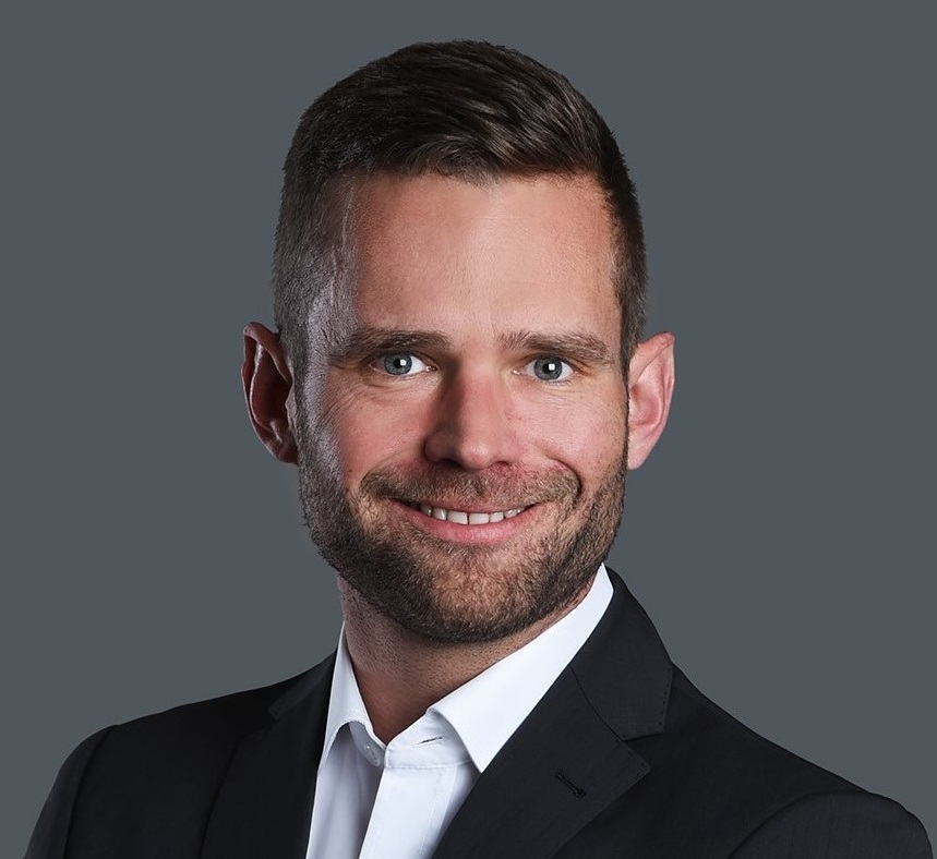 Porträt eines lächelnden Mannes im dunklen Anzug vor grauem Hintergrund; professionelles Business-Foto für Unternehmenswebseiten.