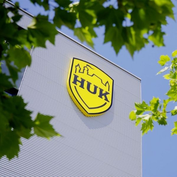 Logo HUK-COBURG su una facciata moderna, incorniciato da chiome verdi sotto un cielo azzurro.