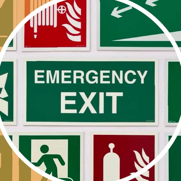 Sammlung von Sicherheitsschildern, darunter ein großes grünes Schild mit der Aufschrift „Emergency Exit“, umgeben von Piktogrammen für Fluchtwege, Feuerlöscher und Notausgänge.