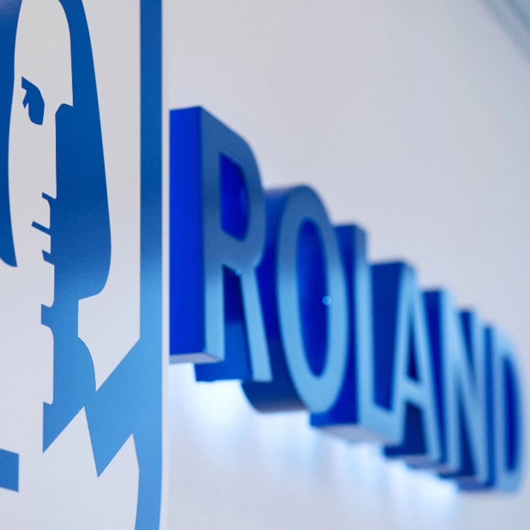 Nahaufnahme des Logos von ROLAND Rechtsschutz, einem Kunden von BSI Software. Der blaue, dreidimensionale Schriftzug «ROLAND» ist an einer weissen Wand angebracht. Links daneben ist das markante blaue Unternehmenssymbol mit einem stilisierten Gesicht zu sehen. Die perspektivische Aufnahme hebt das Logo und die plastische Gestaltung hervor.
