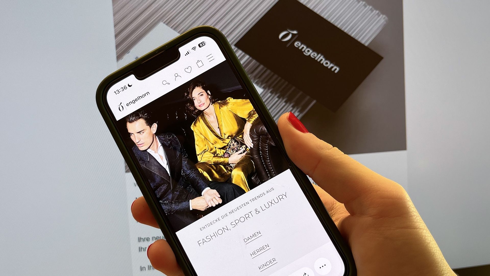 Smartphone in a woman's hand displaying the fashion retailer engelhorn's mobile app with the slogan “Discover the latest trends in Fashion, Sport & Luxury” and an image of stylishly dressed models; a blurred engelhorn loyalty card is visible on a screen in the background.