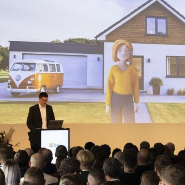 Olav Lenz präsentiert beim Customer Summit 2025 auf einer Bühne vor Publikum die 3D-Kundin Lena Meier vor Einfamilienhaus mit Garage. Die Szene zeigt eine innovative Produktdemonstration und veranschaulicht Customer Relationship Management, digitale Beratung und personalisierte Customer Experience mit der BSI Customer Suite.