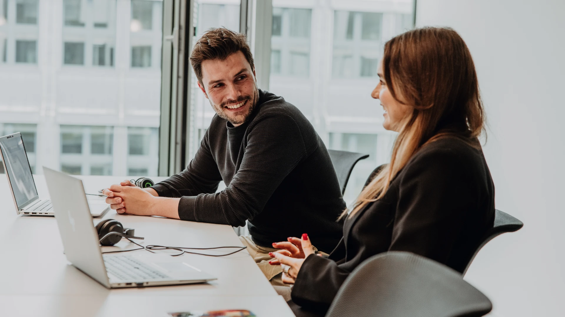 Zwei Mitarbeitende arbeiten gemeinsam an Laptops in einem modernen Büro, tauschen sich lachend aus und zeigen eine offene, kollegiale Arbeitsatmosphäre – authentischer Einblick in die Unternehmenskultur und das Arbeiten bei BSI Software.