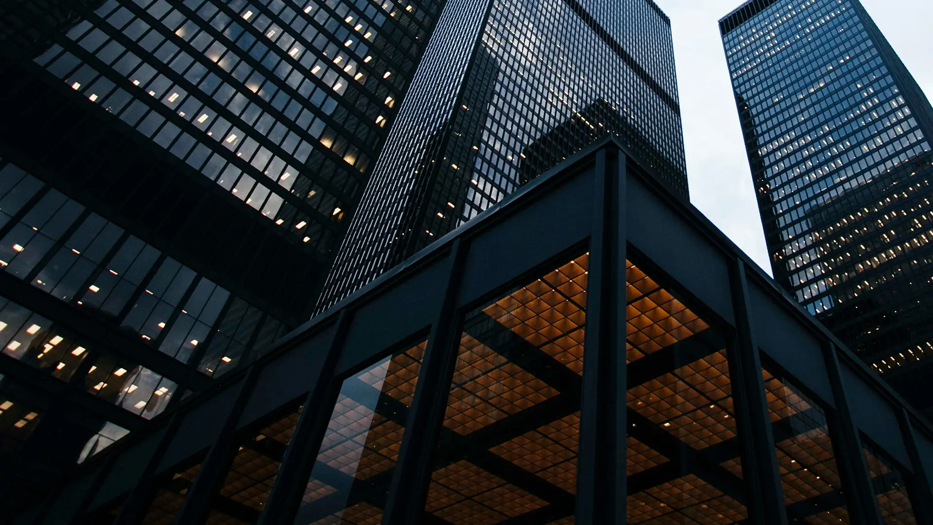 Aus der Froschperspektive aufgenommene, dunkel verglaste Bankengebäude mit beleuchteten Bürofenstern und markantem Eingangsbereich. Die moderne Finanzarchitektur steht sinnbildlich dafür, dass PostFinance und Migros Bank ihre Partnerschaft mit BSI Software ausbauen.