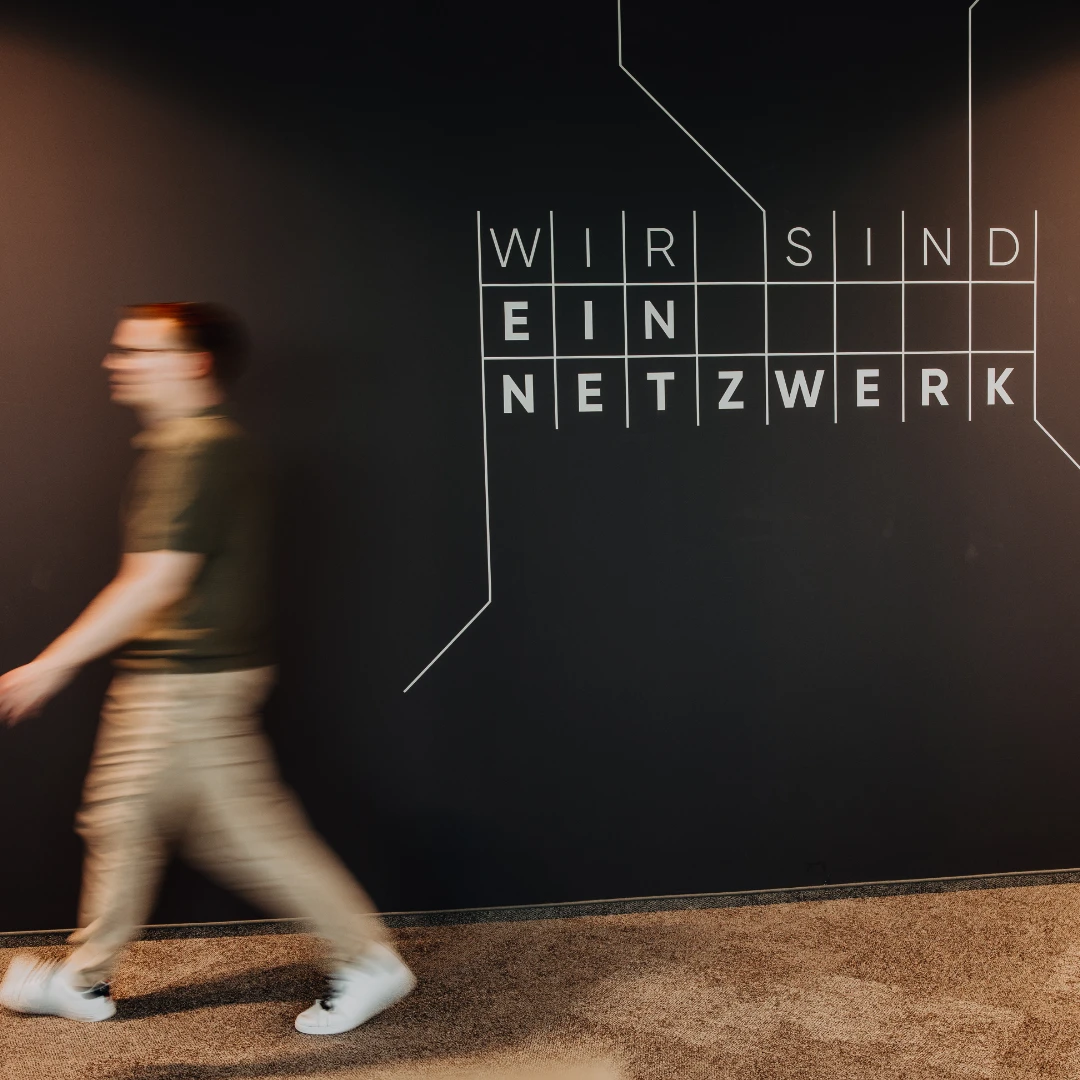 Mitarbeitender geht an einer Wand mit der Aufschrift «Wir sind ein Netzwerk» vorbei – modernes Office-Design und gelebte Vernetzung spiegeln die offene Unternehmenskultur und attraktive Arbeitsumgebung bei BSI Software wider.