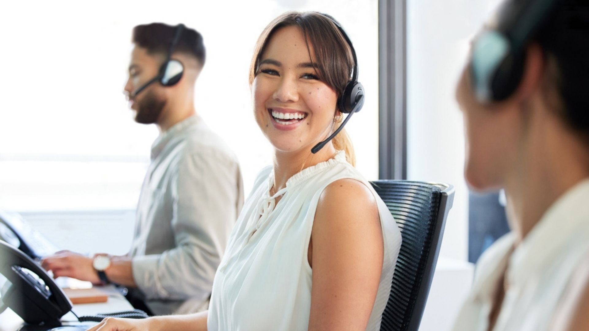Freundliche Callcenter-Mitarbeiterin mit Headset lächelt in die Kamera, während sie mit Kollegen im modernen Büro telefonischen Kundenservice anbietet.