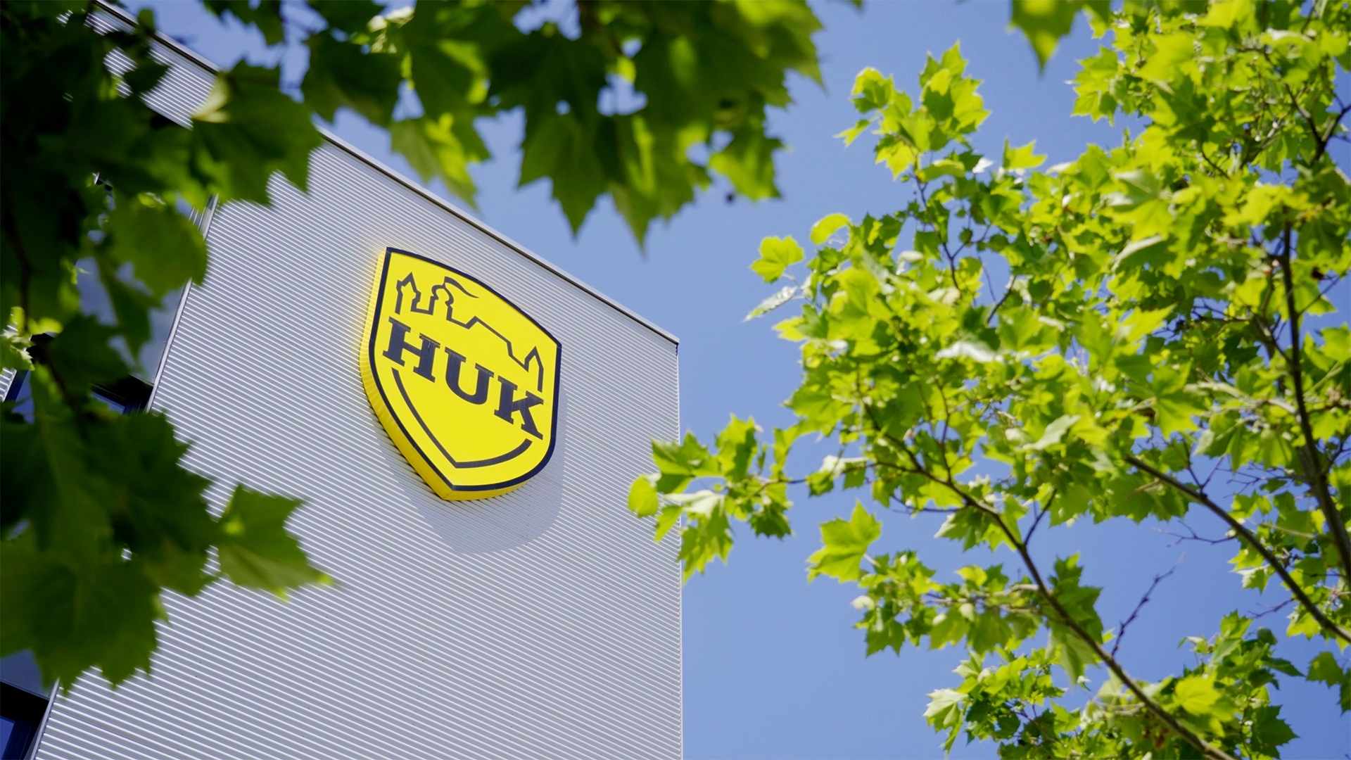 HUK-COBURG Logo an moderner Gebäudefassade, aufgenommen durch grüne Baumkronen vor blauem Himmel.