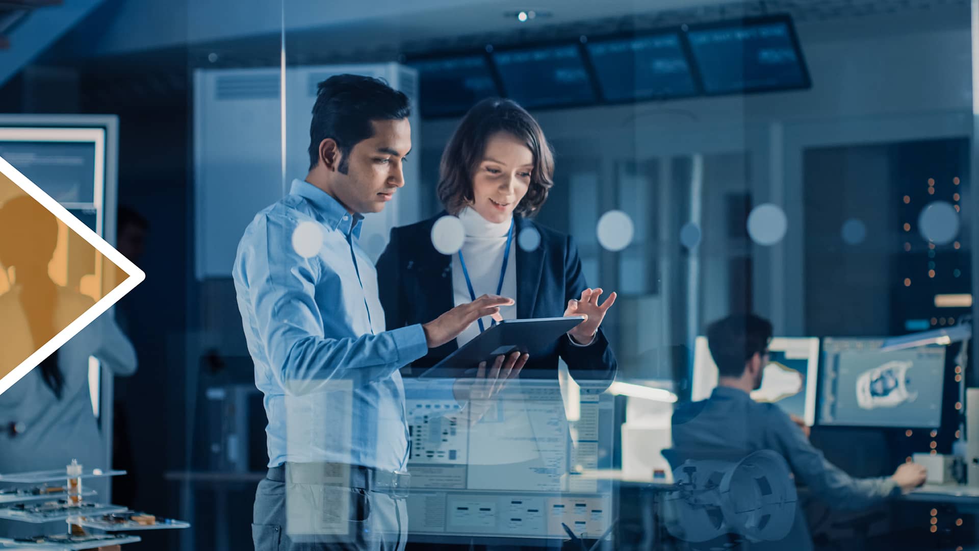 Zwei Fachleute in moderner Büroumgebung analysieren Daten auf einem Tablet. Im Hintergrund sind Computer, Bildschirme und technische Geräte sichtbar – Symbol für digitale Transformation und innovative Teamarbeit.