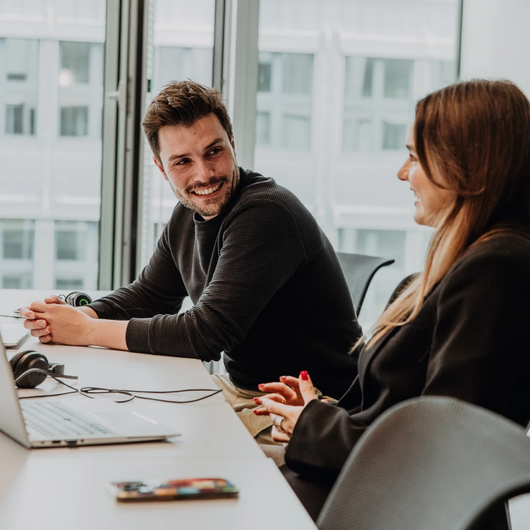 Zwei Mitarbeitende arbeiten gemeinsam an Laptops in einem modernen Büro, tauschen sich lachend aus und zeigen eine offene, kollegiale Arbeitsatmosphäre – authentischer Einblick in die Unternehmenskultur und das Arbeiten bei BSI Software.